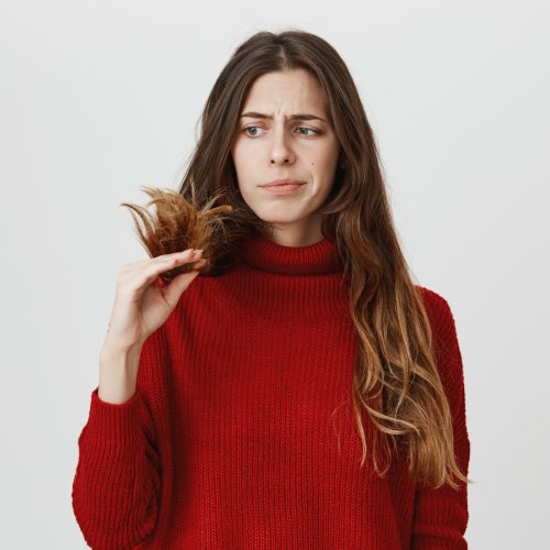 Dissatisfied young woman in casual clothes looking at her dark long hair, being displeased with work of hairdresser, frowning her face in discontent. Negative emotions and face expression